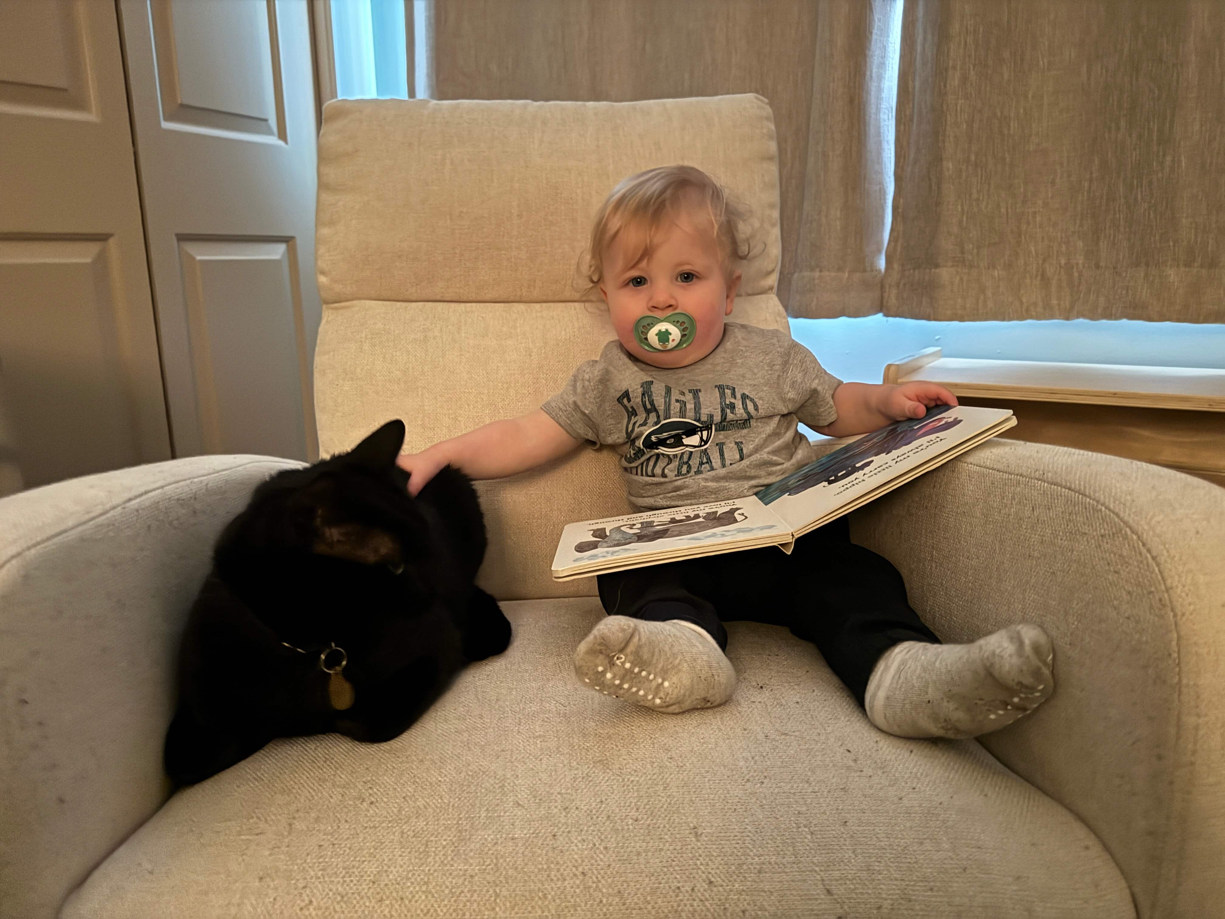 a baby looking at a book and petting a black cat (aw)